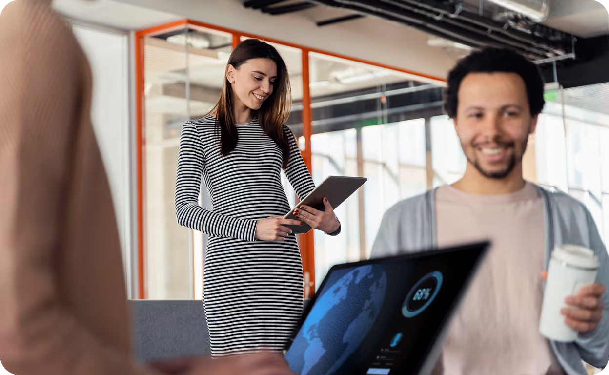 Smiling woman in striped dress holding a tablet and a man holding a coffee cup near a laptop displaying a world map and data in a modern office.