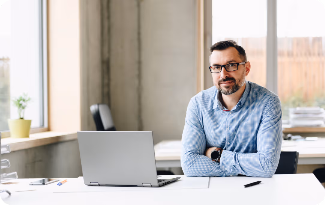 Man with glasses and beard sitting at a desk with a laptop in a bright office.
