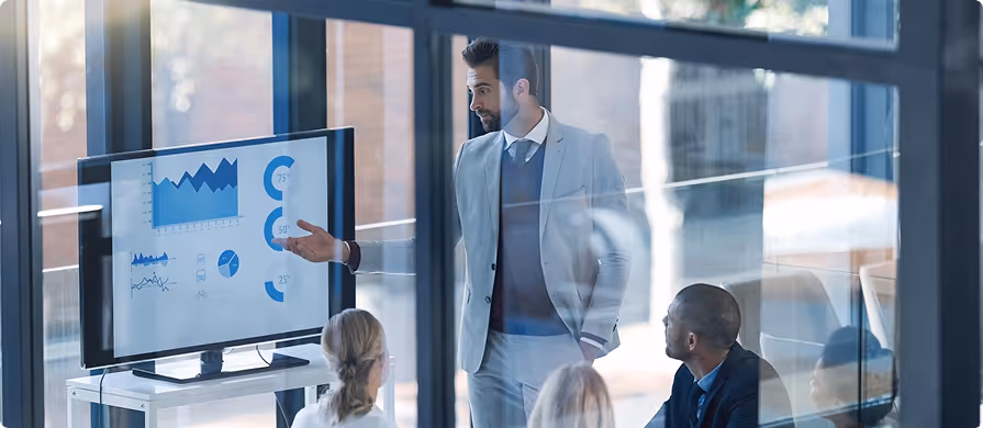 Businessman in a suit presenting data charts on a screen to colleagues in a modern office.