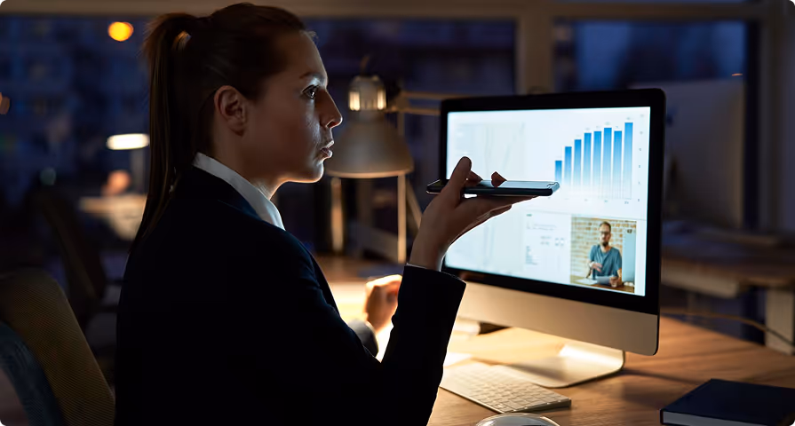 Woman in a dimly lit office holding a smartphone near her mouth while looking at a computer screen showing charts and a video call.
