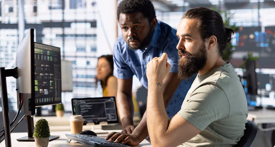 Two men collaborating and looking at a computer screen in a modern office with a woman working in the background.