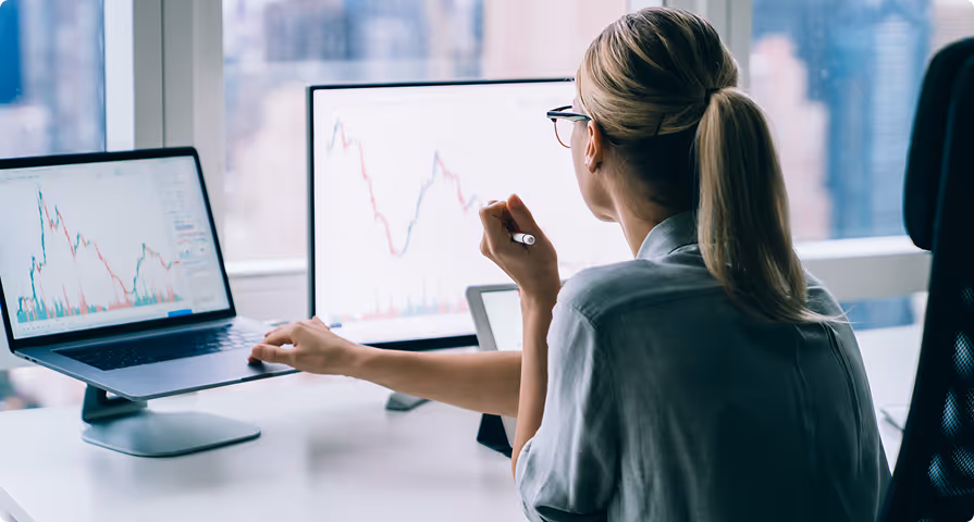 Woman working with financial charts on a laptop and a desktop monitor in a modern office with a city view.