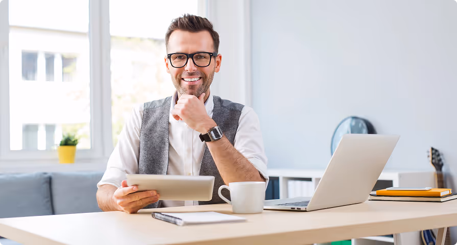 Smiling man wearing glasses and a vest sitting at a desk with a tablet, laptop, coffee cup, and notebook in a bright office.