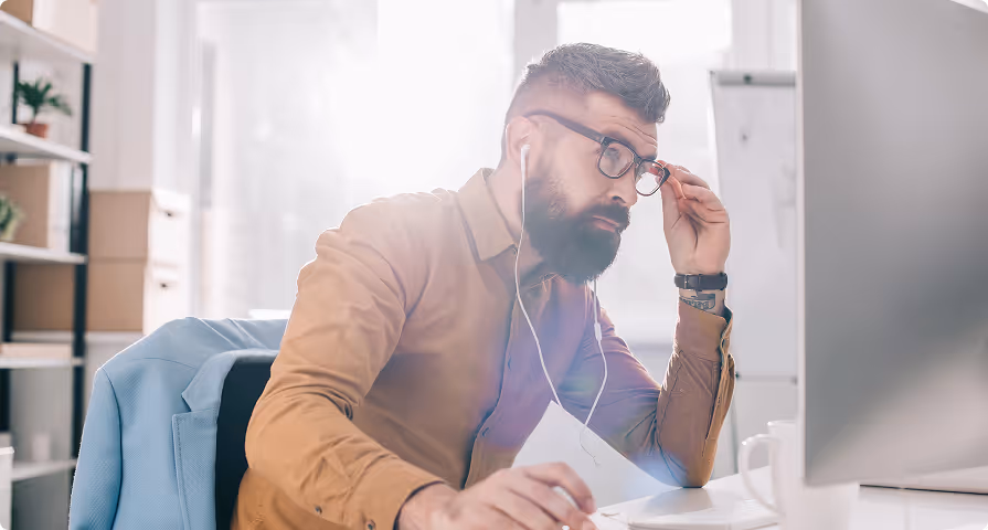 Bearded man wearing glasses and earphones, adjusting his glasses while working on a computer in a bright office.
